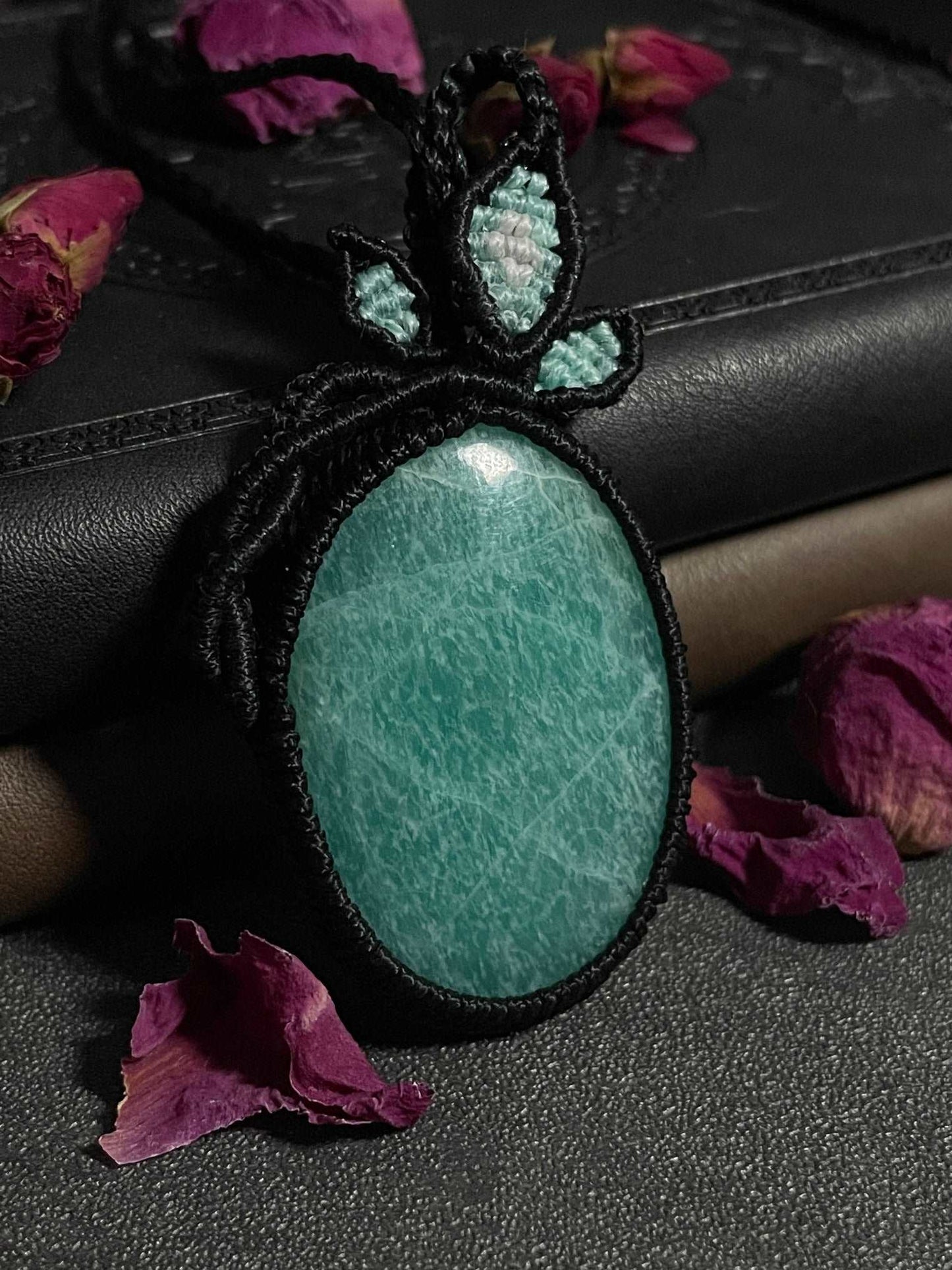 Pictured is an amazonite cabochon wrapped in macrame thread. A gothic book and flowers are nearby. Amazonite Macramé Necklace (Twisted Nightshade Jewellery) - The Wandering Fox Emporium, Your Crystal Store close up 2