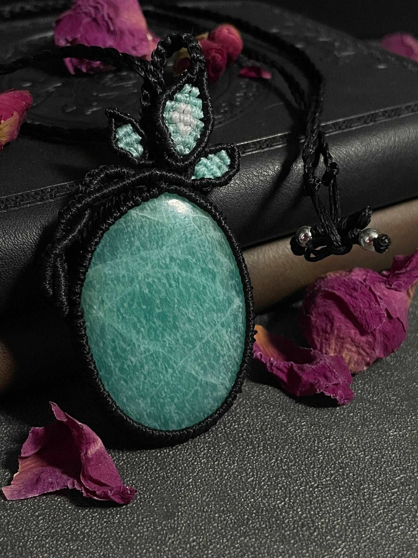 Pictured is an amazonite cabochon wrapped in macrame thread. A gothic book and flowers are nearby. Amazonite Macramé Necklace (Twisted Nightshade Jewellery) - The Wandering Fox Emporium, Your Crystal Store close up