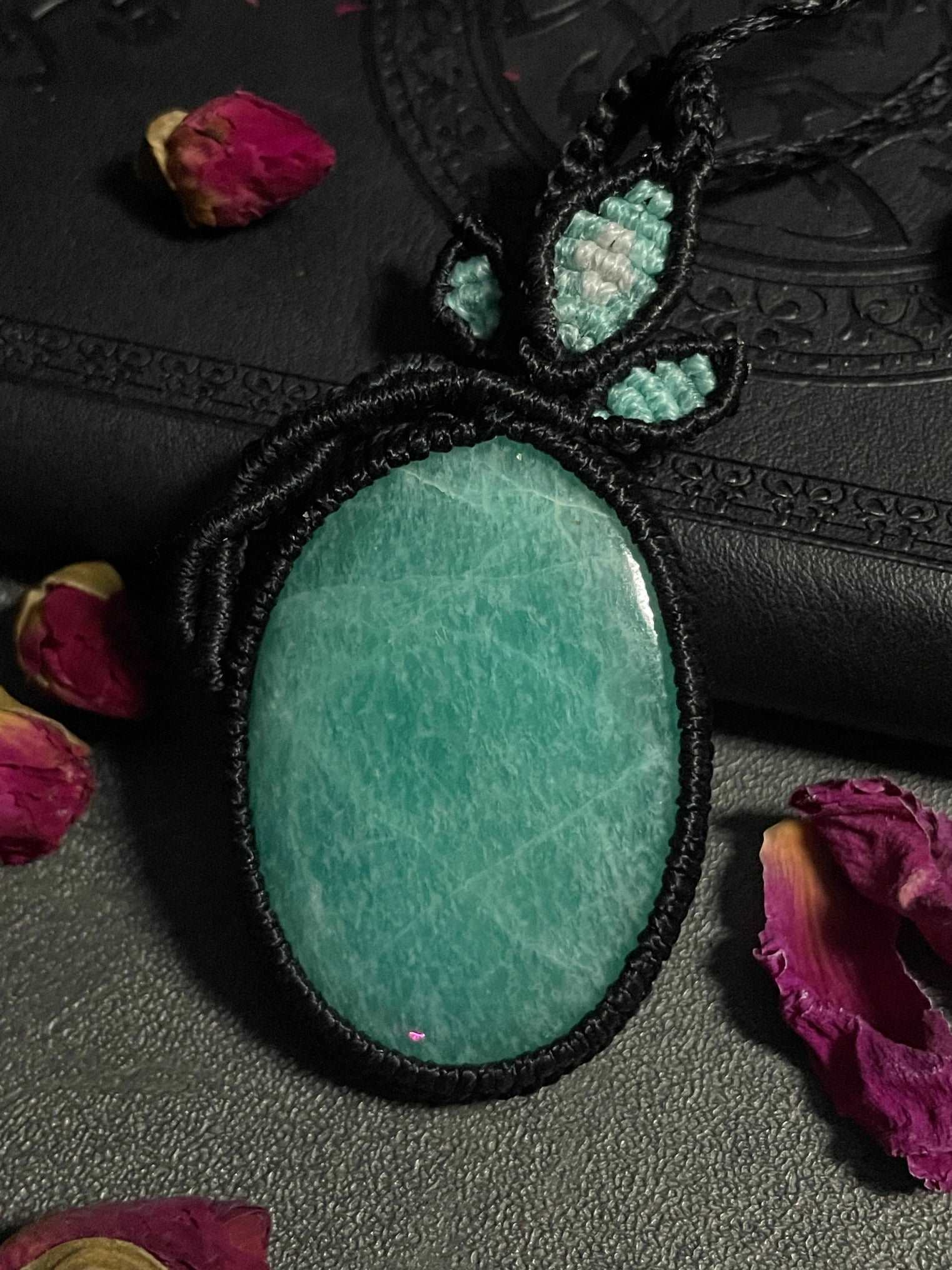 Pictured is an amazonite cabochon wrapped in macrame thread. A gothic book and flowers are nearby. Amazonite Macramé Necklace (Twisted Nightshade Jewellery) - The Wandering Fox Emporium, Your Crystal Store front
