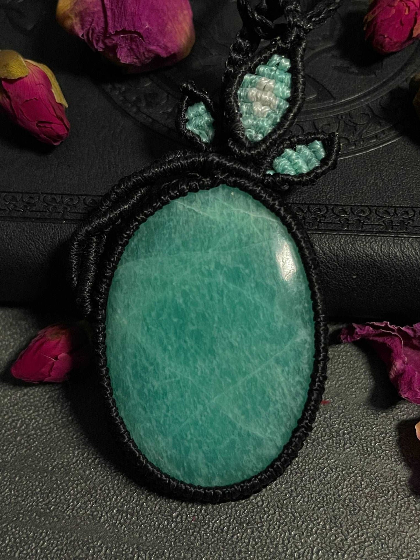 Pictured is an amazonite cabochon wrapped in macrame thread. A gothic book and flowers are nearby. Amazonite Macramé Necklace (Twisted Nightshade Jewellery) - The Wandering Fox Emporium, Your Crystal Store
