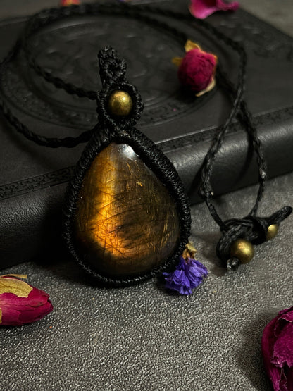 Pictured is a labradorite cabochon wrapped in macrame thread. A gothic book and flowers are nearby. Sunset Labradorite Teardrop Macramé Necklace (Twisted Nightshade Jewellery) - The Wandering Fox Emporium, Your Crystal Store